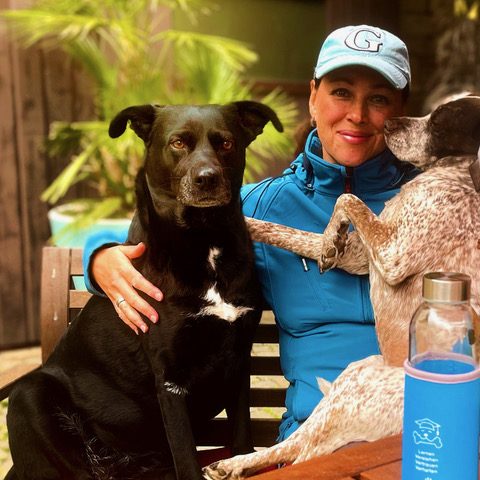 Miri Styppa, Tierpsychologin und Hundetrainerin Pfötchenhof Pfalz Frau mit schwarzem Hund und gestromtem Hund auf einem Holzstuhl in einem Garten.